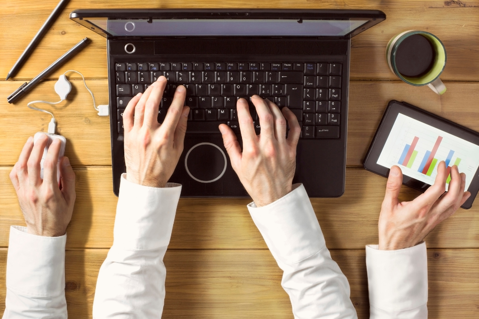 An aerial view of a man multitasking with hands on the laptop and two extra hands, one holding a tablet demonstrating