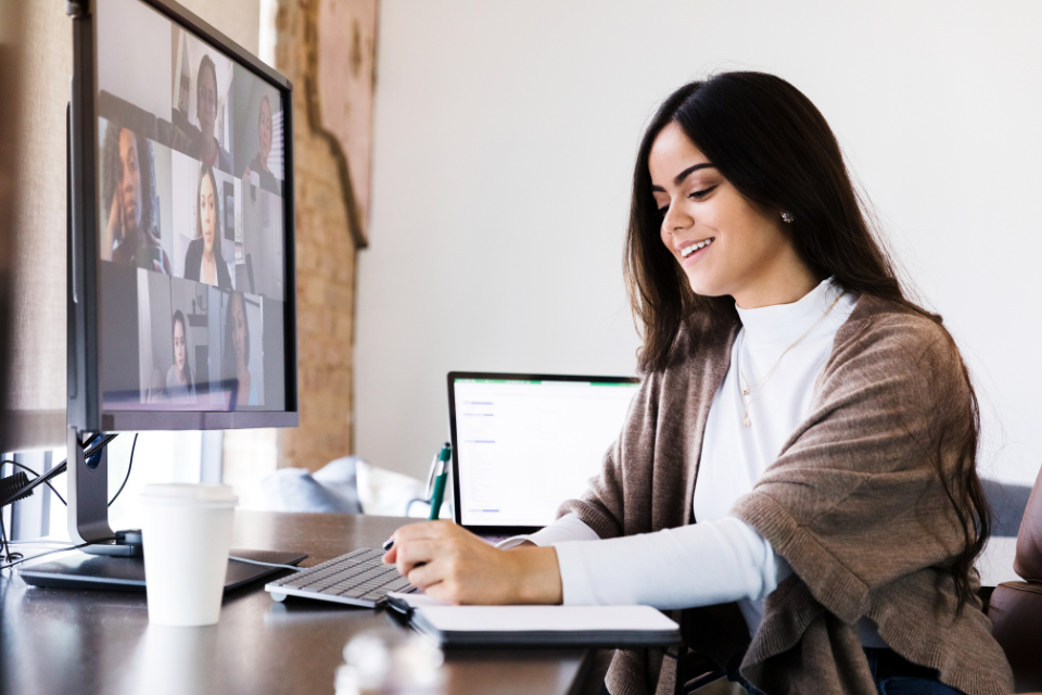 A woman at her desk on a Zoom call takes notes on paper