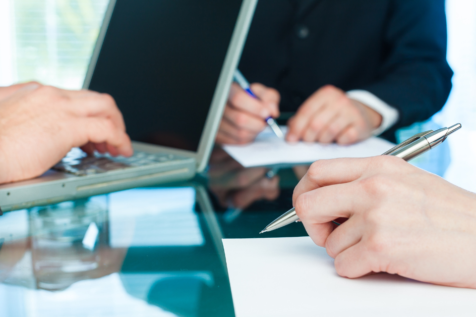 Two people taking meeting notes with pen and paper and one person taking notes on a laptop