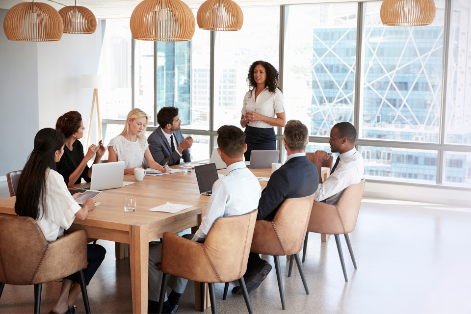 Six people sit at a table during and one stands in front for a work meeting