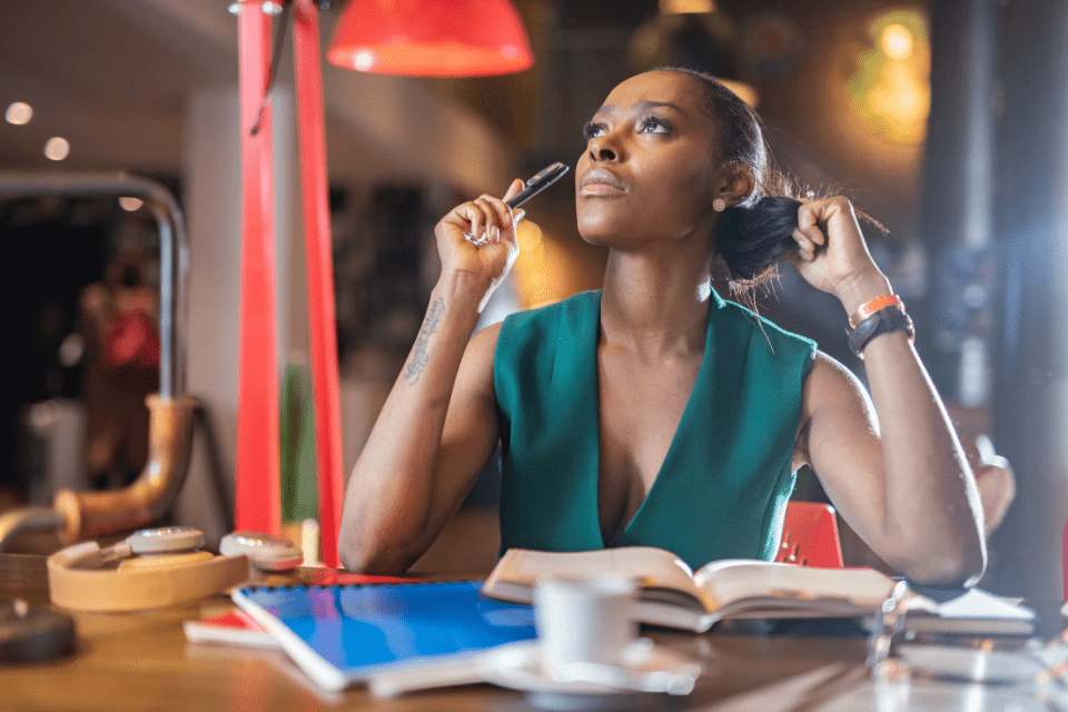 A woman sits at a desk with an open book and looks up, thinking