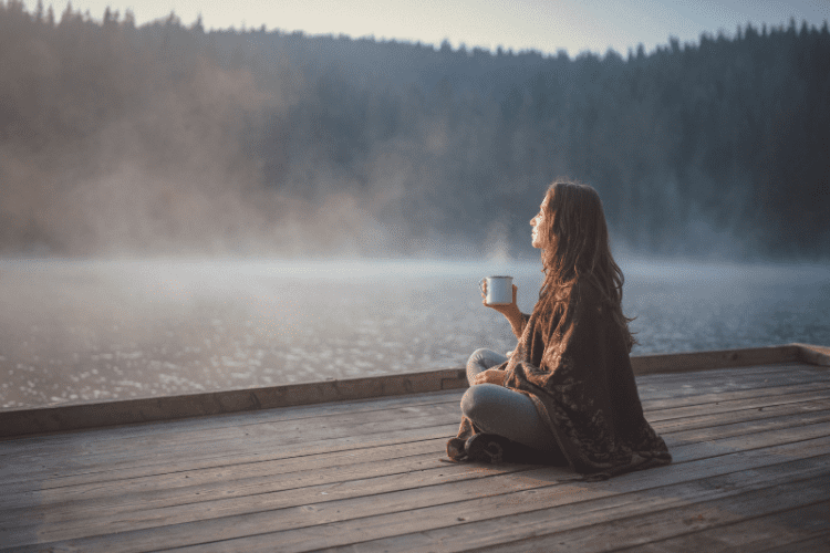 A woman sits on a pier overlooking a lake covered with fog and enjoys coffee