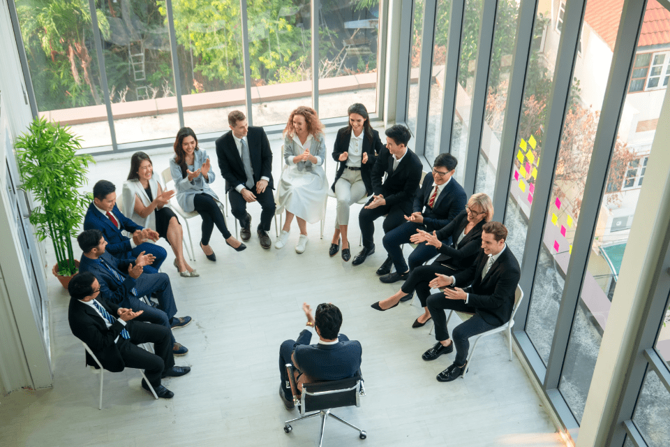 A corporate trainer talks to a team of 15 who are seated in a semi-circle in a conference room