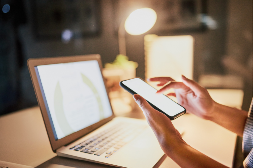 A person in front of an open laptop checks their iphone