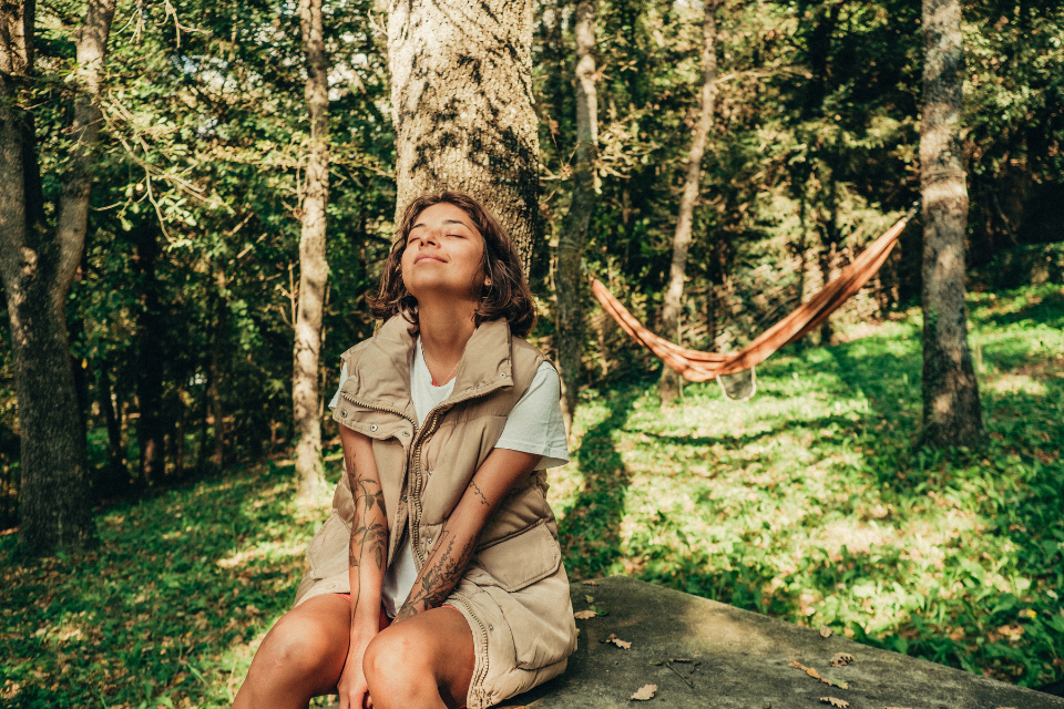 A woman sits in yard under trees with eyes closed, face to the sun, daydreaming in nature.