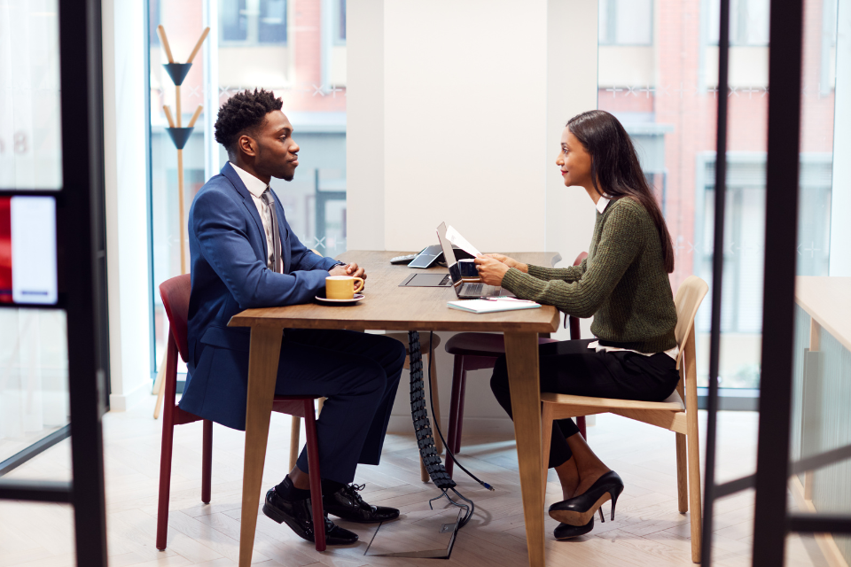 A man and woman sit across from each other at a table. Both are dressed professionally.