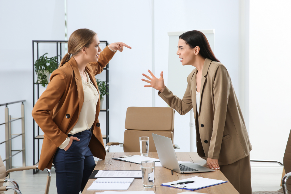 Two women stand on either side of a desk at work arguing