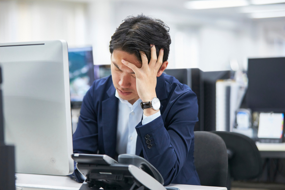 Man sits in front of computer monitor at office with hand on his forehead. He is stressed.