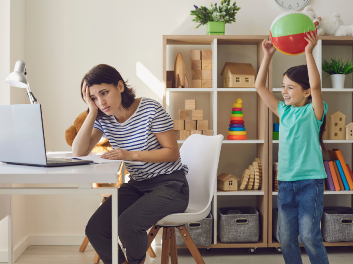 A working mom tries to focus on her computer while a young child stands behind her holding a beach ball