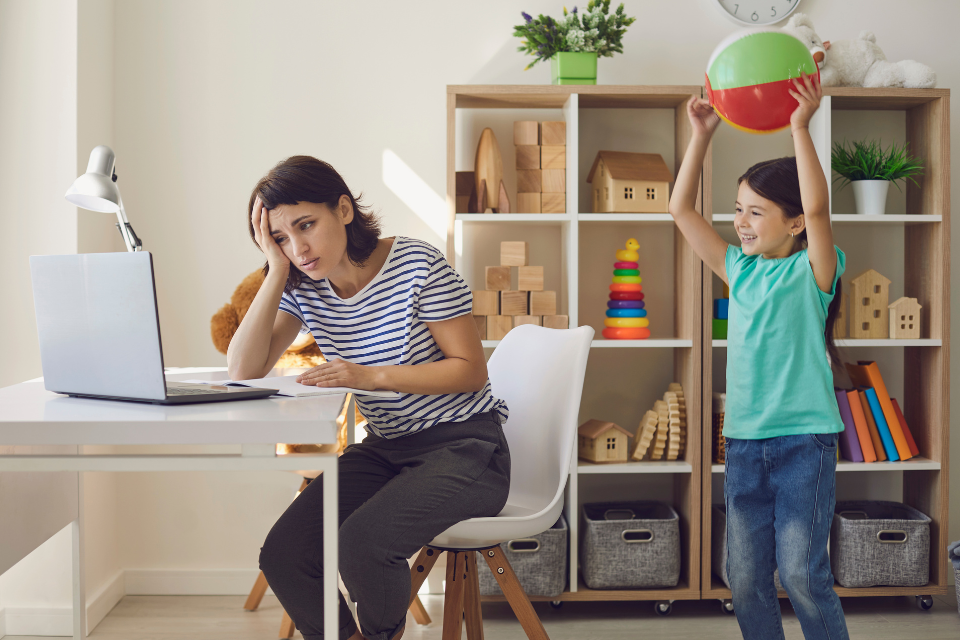 A working mom tries to focus on her computer while a young child stands behind her holding a beach ball