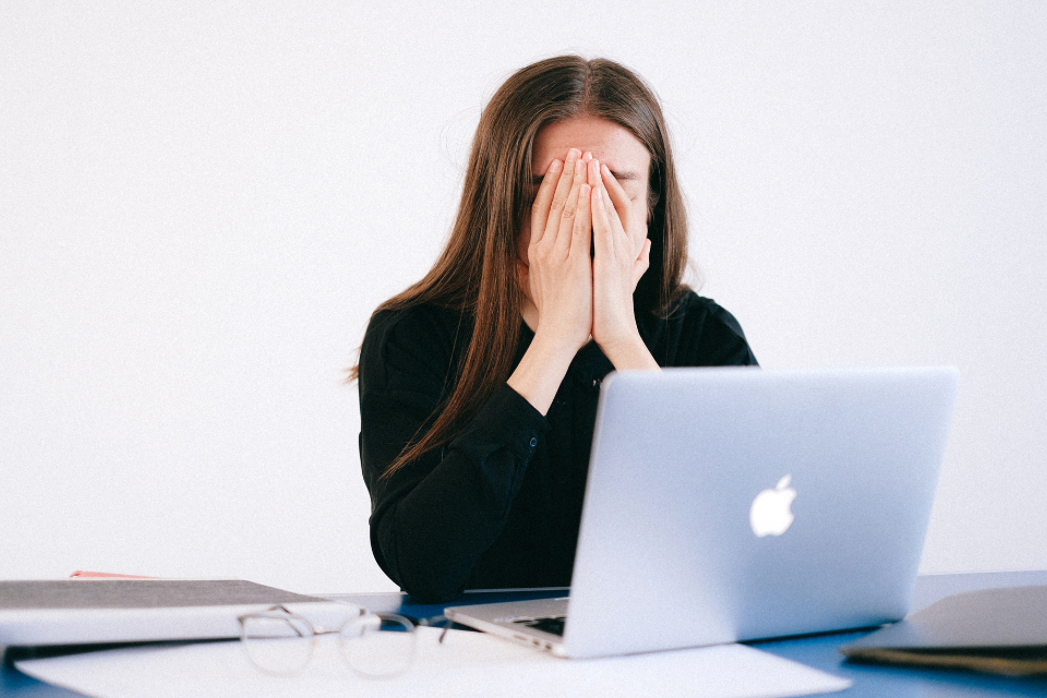A woman in business suit with hands covering her face. She sits in front of a laptop