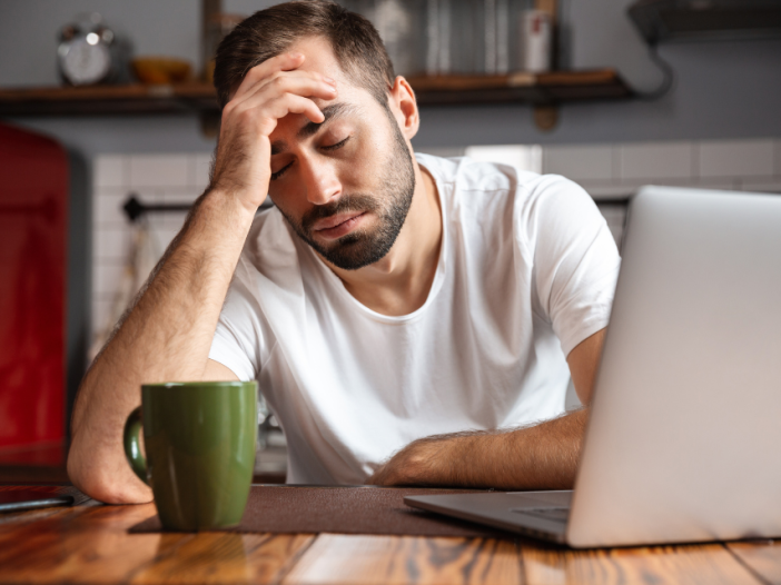 Man sits at kitchen table with hand at forehead in front of laptop. There's a coffee cup in front of him. He's exhausted and overworked