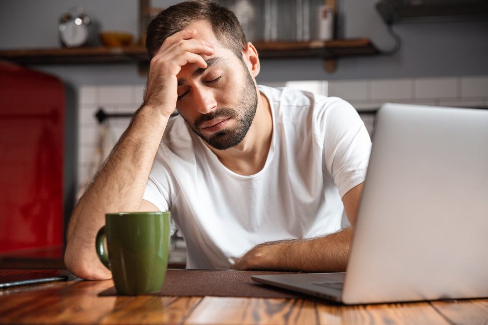 Man sits at kitchen table with hand at forehead in front of laptop. There's a coffee cup in front of him. He's exhausted and overworked