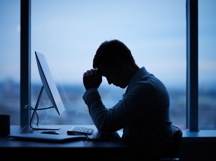 A man whose productivity is going down is in silhouette sits at his desk, his head in his hand, next to a window after sundown