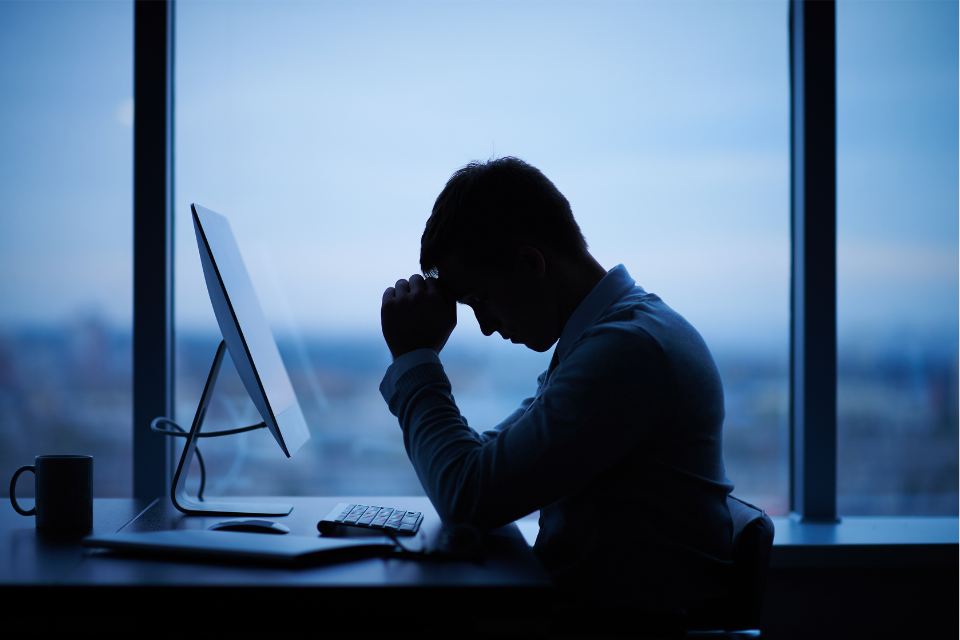A man whose productivity is going down is in silhouette sits at his desk, his head in his hand, next to a window after sundown